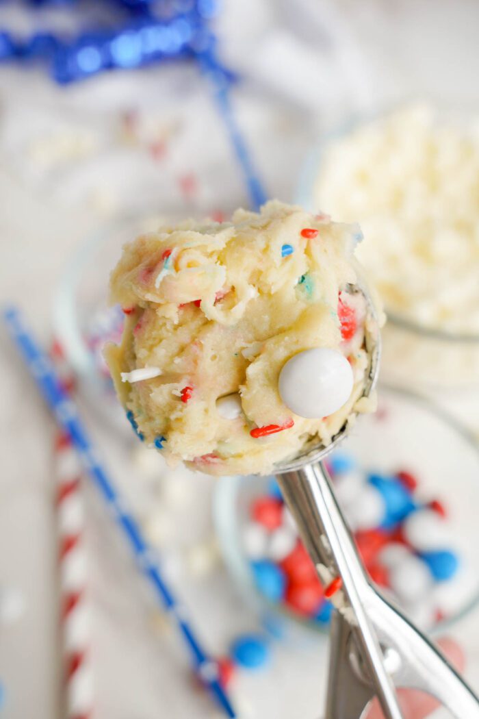 Scoop of birthday cake cookie dough with sprinkles and a white chocolate ball, held over a bowl in a festive 4th of July setting.