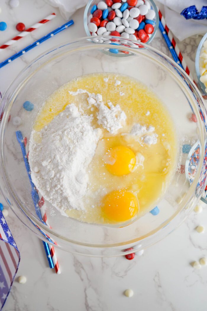 Glass bowl of baking ingredients with eggs, flour, and butter, surrounded by colorful candies and patriotic decorations for easy 4th of July cake mix cookies.