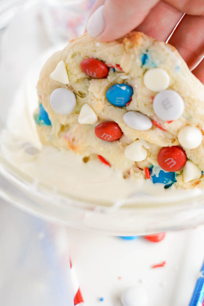 A hand holding a freshly baked cookie with colorful candy pieces and white chocolate chips, over a bowl of cookie dough, resembling an easy 4th of July celebration treat.
