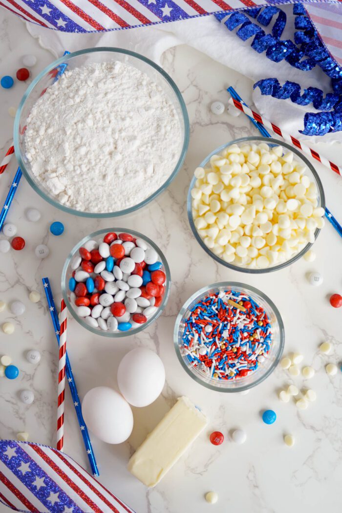 Overhead view of baking ingredients on a marble surface, including flour, eggs, butter, M&Ms, white chocolate chips, and sprinkles for Easy 4th of July Cake Mix Cookies.