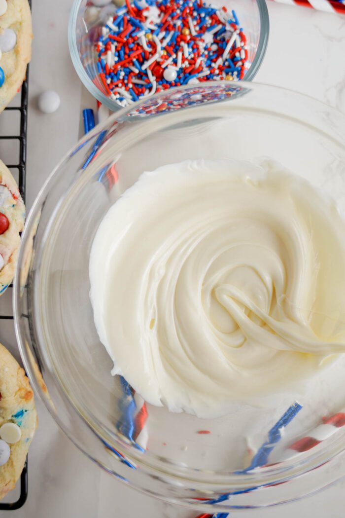 Frosting being swirled in a glass bowl with Easy 4th of July Cake Mix Cookies and red, white, and blue sprinkles in the background.