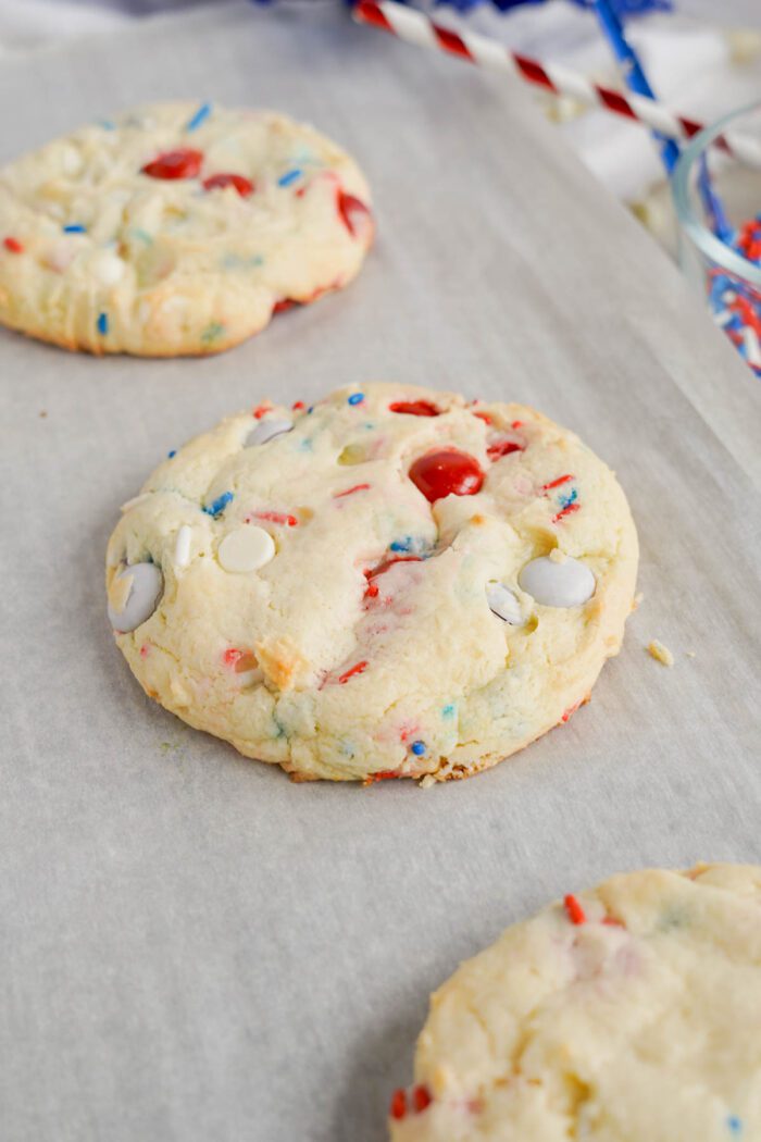 Freshly baked Easy 4th of July Cake Mix Cookies with colorful candy pieces on parchment paper, with blurred holiday-themed decorations in the background.