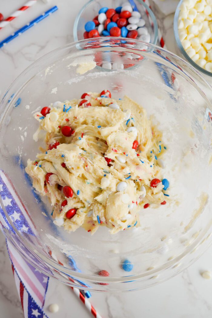 Cookie dough in a glass bowl with red, white, and blue candies mixed in for Easy 4th of July Cake Mix Cookies, on a marble countertop with additional candies and decoration visible.