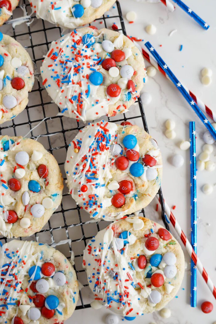 Easy 4th of July cake mix cookies with colorful candy coverings, white frosting, and sprinkles on a cooling rack, surrounded by festive straws and candy pieces.