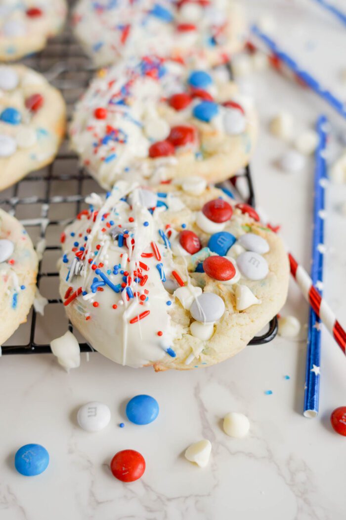 Easy homemade cookies with white icing and patriotic red, white, and blue sprinkles, cooling on a wire rack with extra candies scattered around.