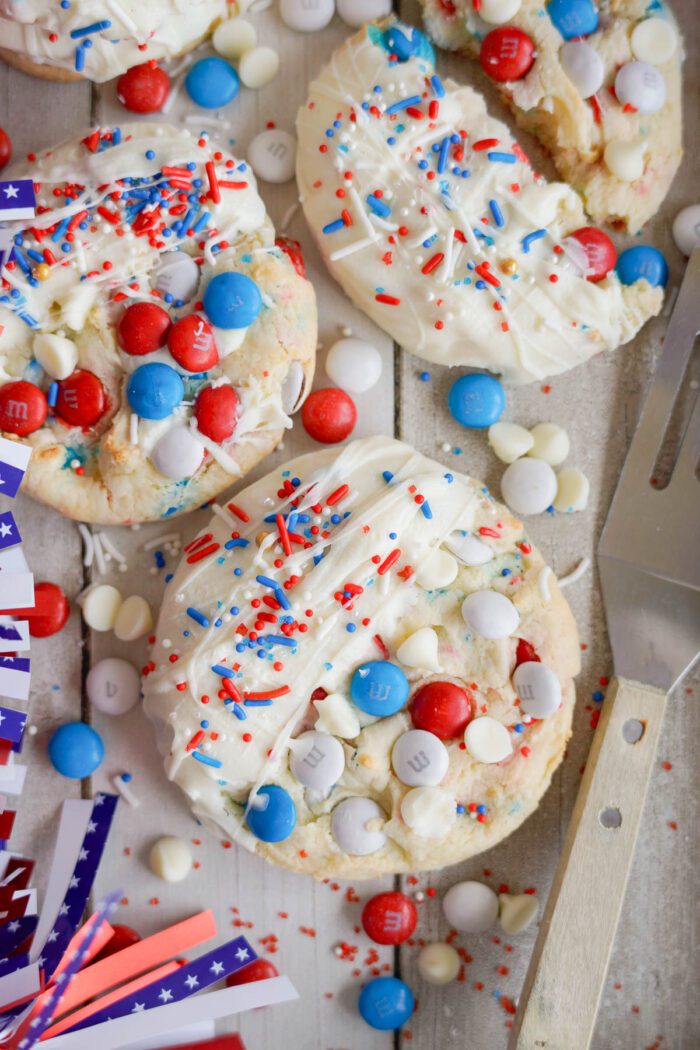 Easy 4th of July Cake Mix Cookies with white frosting, red and blue candies, and sprinkles on a surface with small American flags.