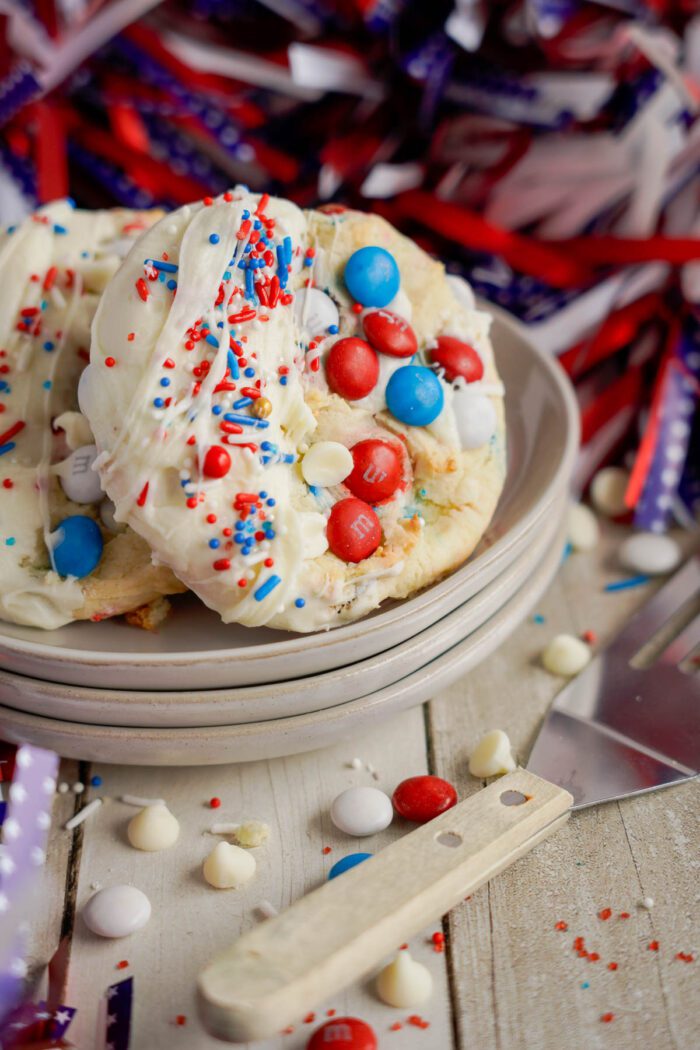 A stack of white chocolate-dipped Easy 4th of July Cake Mix Cookies with red and blue candies, surrounded by festive ribbons on a wooden table.