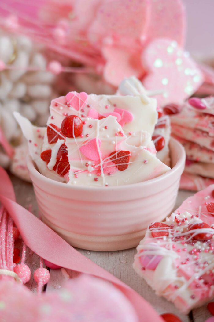 A bowl of white chocolate bark with pink accents and candy pieces, surrounded by ribbons and more sweets on a wooden surface.