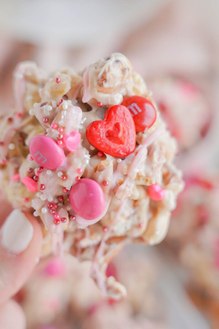 A hand holding a white chocolate treat adorned with pink and red candies and sprinkles.