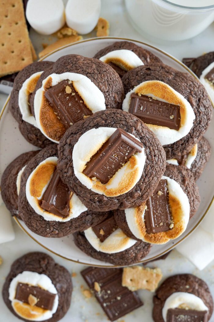 Plate of s'mores cookies with marshmallows and chocolate pieces on top, accompanied by milk and ingredients in the background.