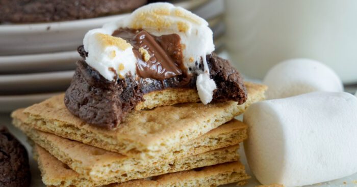 A close-up of a s'more Cookie with a bite taken out, revealing melted chocolate and marshmallow, stacked on graham crackers, with whole marshmallows to the side.