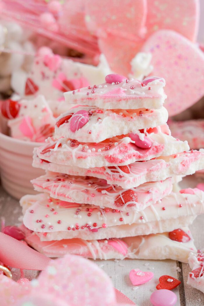 Stack of pink and white valentine's day themed bark with sprinkles and heart decorations, alongside additional treats and pink decorations.