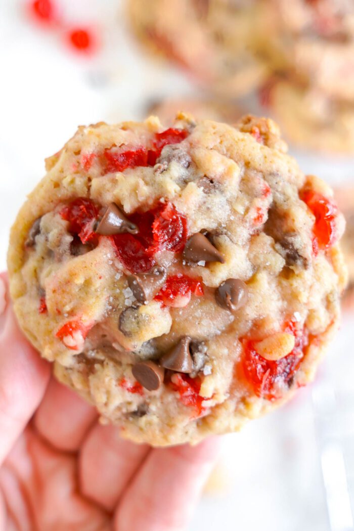 A close-up of a hand holding a Cherry Chocolate Chip cookie with candy pieces.