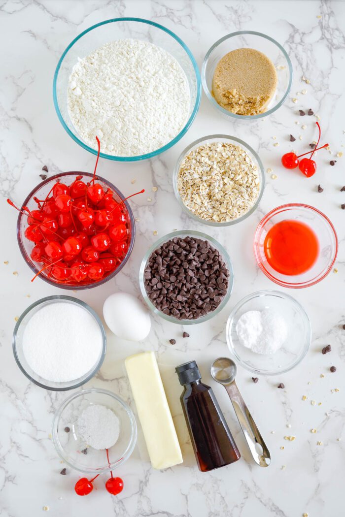 Various baking ingredients for a Cherry Chocolate Chip Cookies recipe neatly arranged on a marble countertop ready for use.