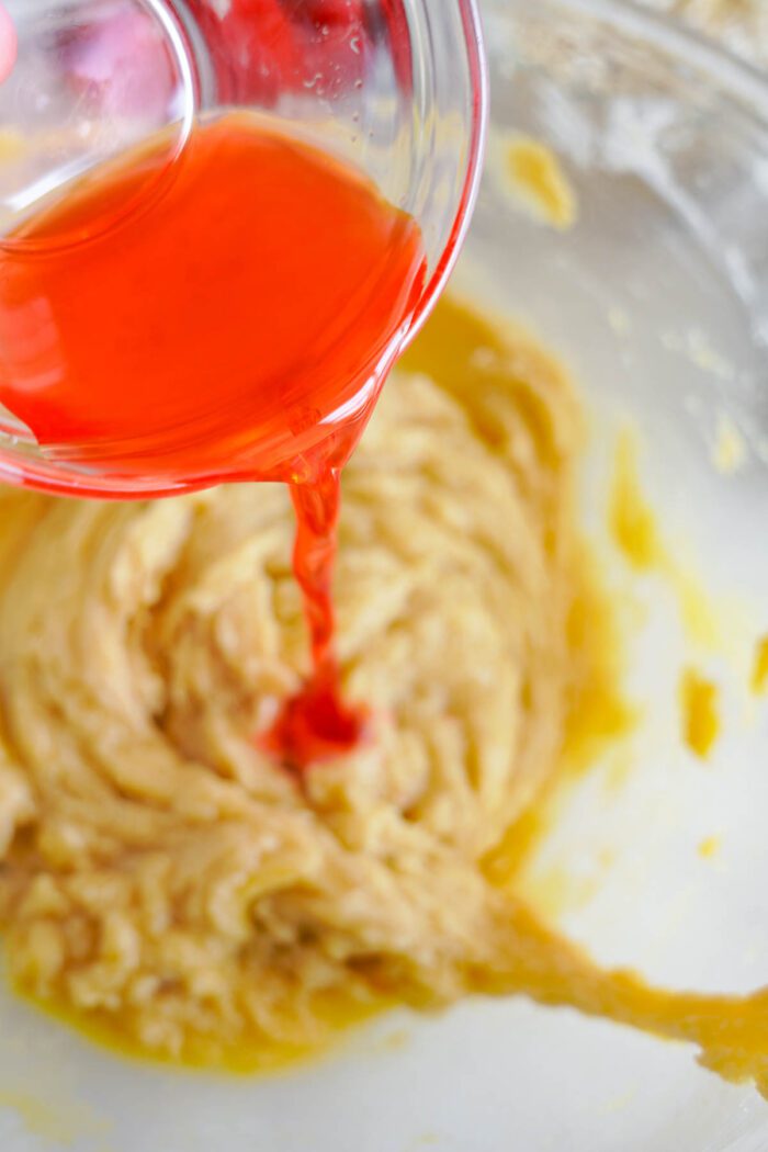 Pouring red food coloring into cake batter for a Cherry Chocolate Chip Cookies recipe in a glass mixing bowl.