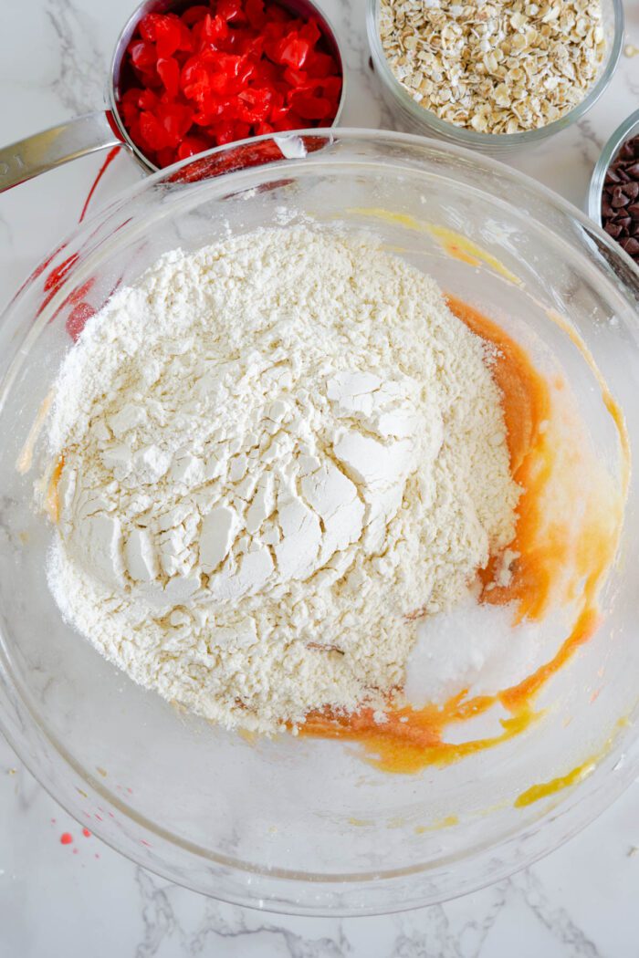 Ingredients for baking in a glass bowl on a marble countertop, including flour, oats, and an egg mixture for Cherry Chocolate Chip Cookies.