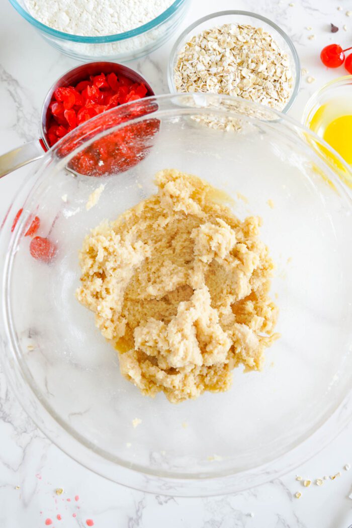 Cookie dough in a glass bowl surrounded by ingredients such as oats, flour, candy sprinkles, and cherry chocolate chips on a marble countertop for a Cherry Chocolate Chip Cookies recipe.