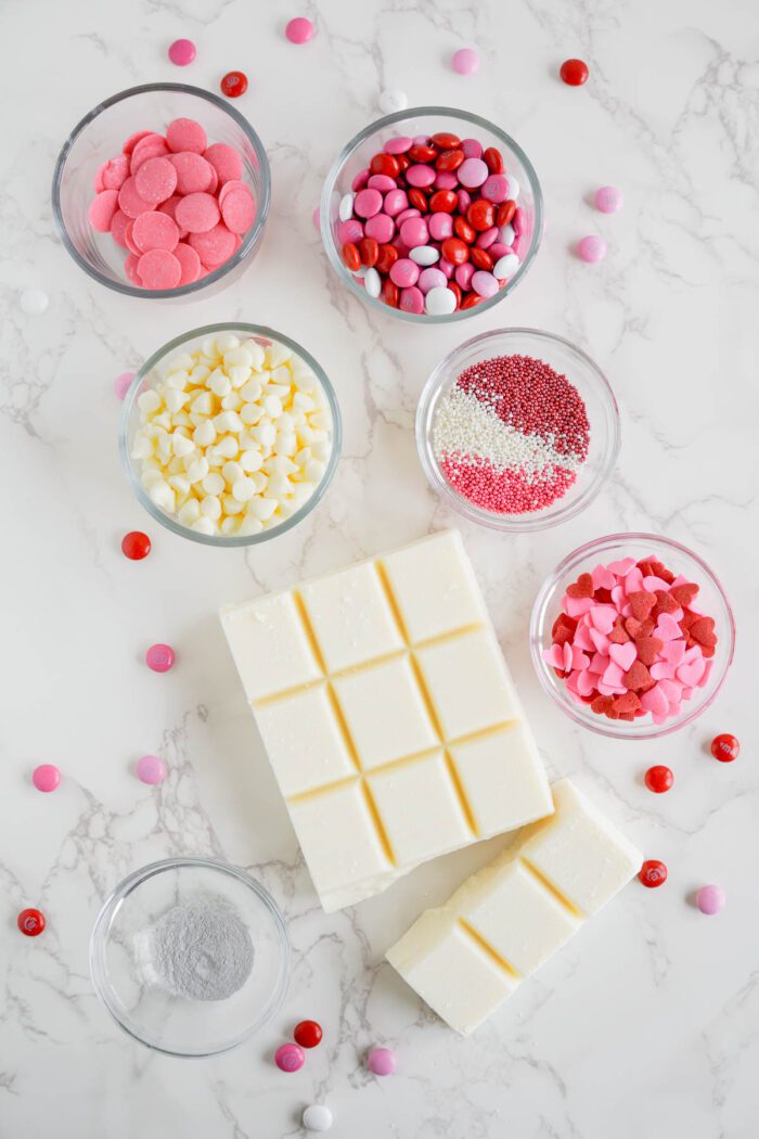 Assorted baking ingredients on a marble countertop, including white chocolate bars and bowls of colorful candy and sprinkles.