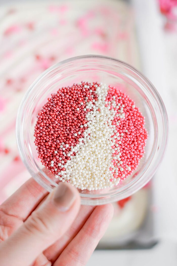 A hand holding a clear glass jar filled with red, pink, and white Valentine's Day sprinkles.