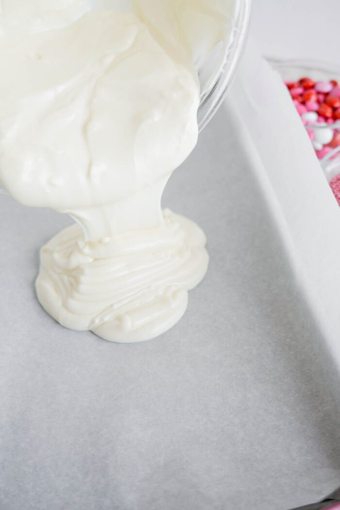 Pouring melted white chocolate from a container onto parchment paper with a box of colorful sprinkles visible in the background.