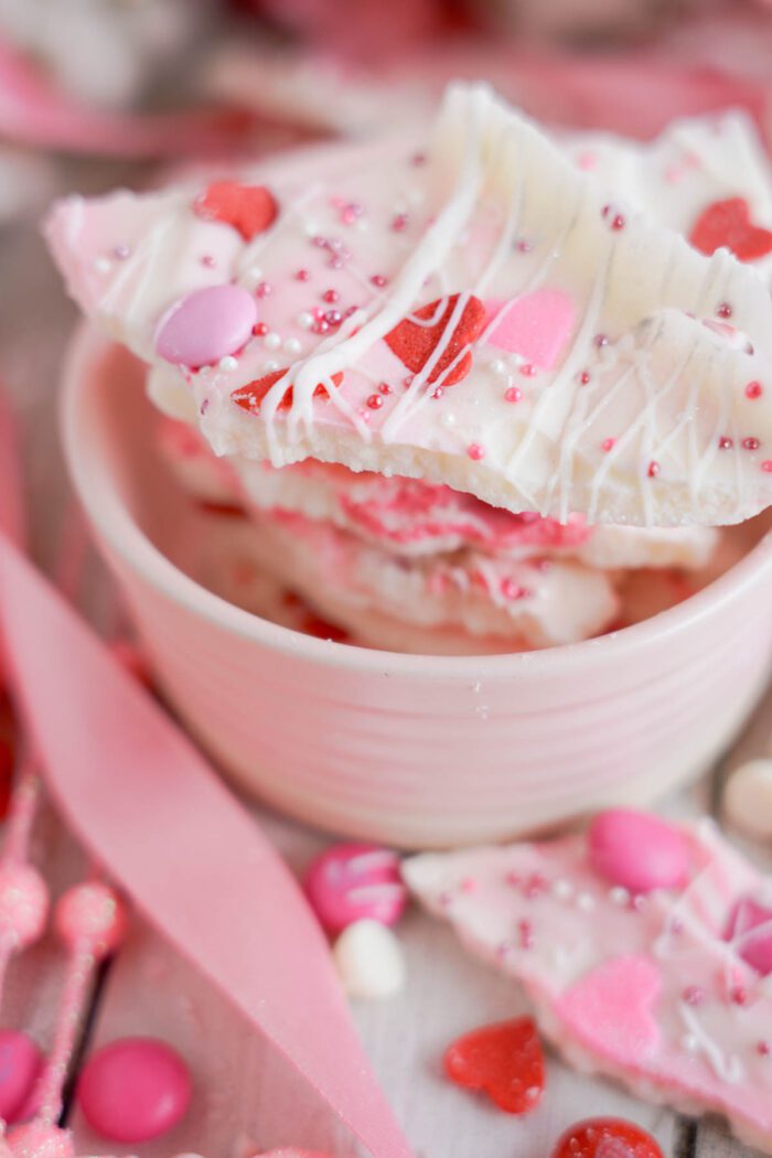 Pink and white valentine's day candy bark in a light pink bowl, garnished with sprinkles and heart-shaped candies.