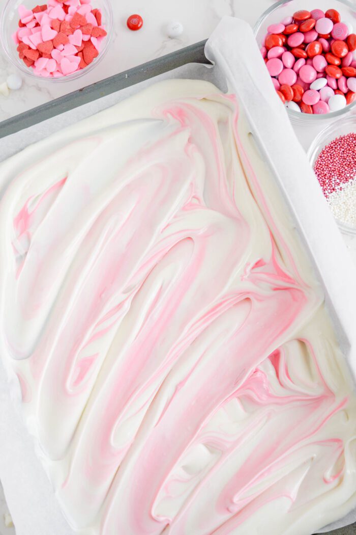 A baking tray filled with pink and white chocolate swirled surrounded by various colorful candies and sprinkles.
