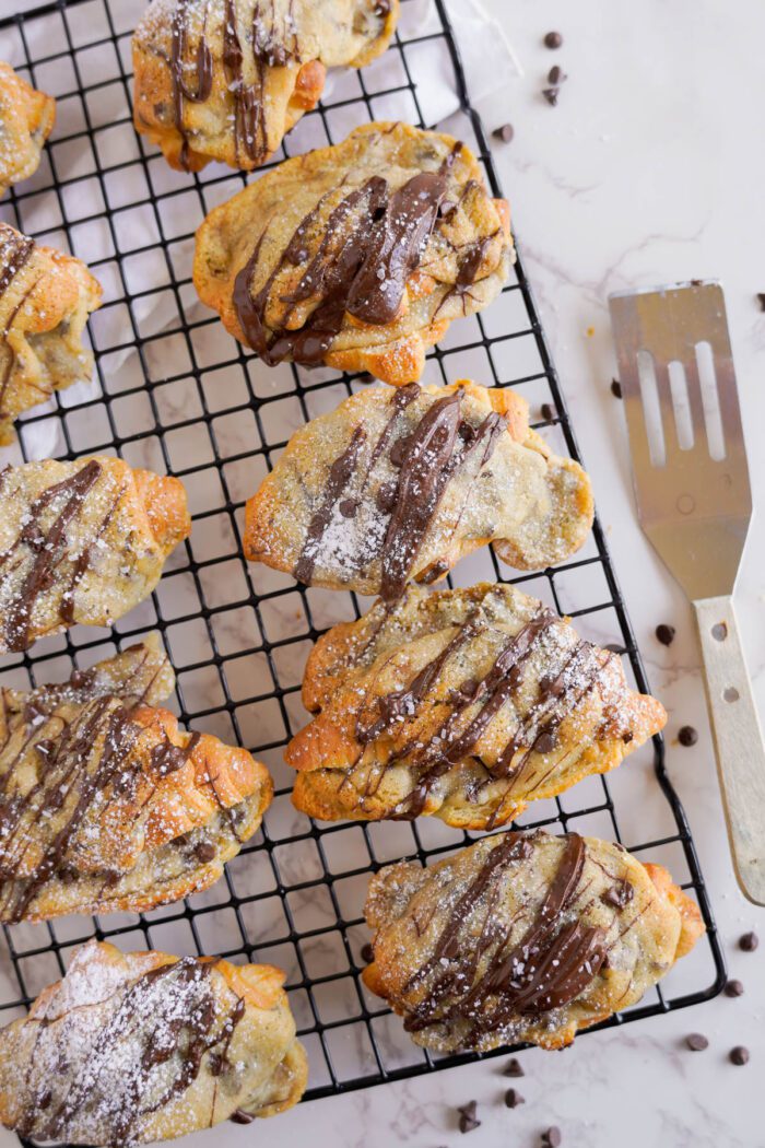 Freshly baked chocolate chip cookie croissants on a cooling rack, sprinkled with powdered sugar, with a spatula nearby.
