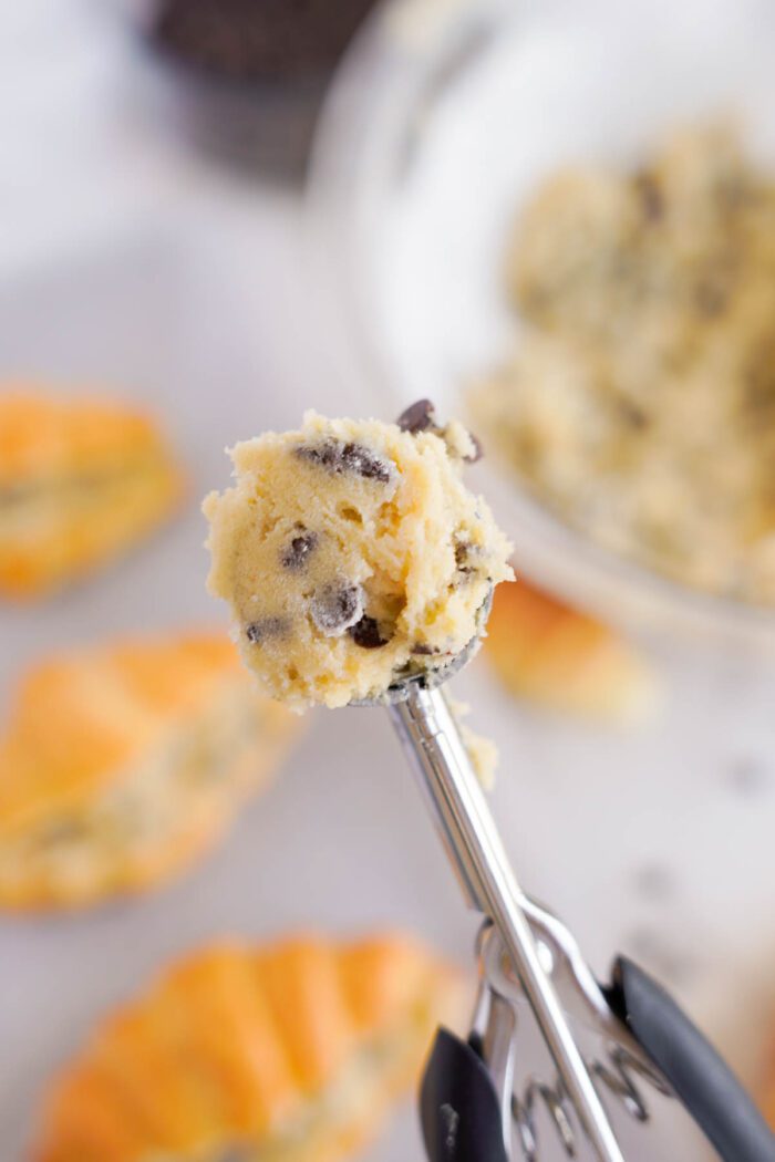 Cookie dough in a scoop with chocolate chips, against a blurred background of more dough and cookies on a white surface, ready for a chocolate chip cookie recipe.
