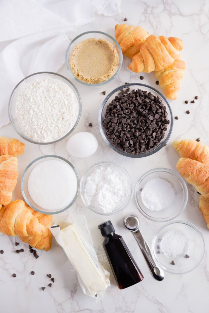 Ingredients for baking laid out on a marble countertop, including flour, sugar, butter, eggs, vanilla, and chocolate chips, with a Chocolate Chip Cookie Croissant recipe nearby.