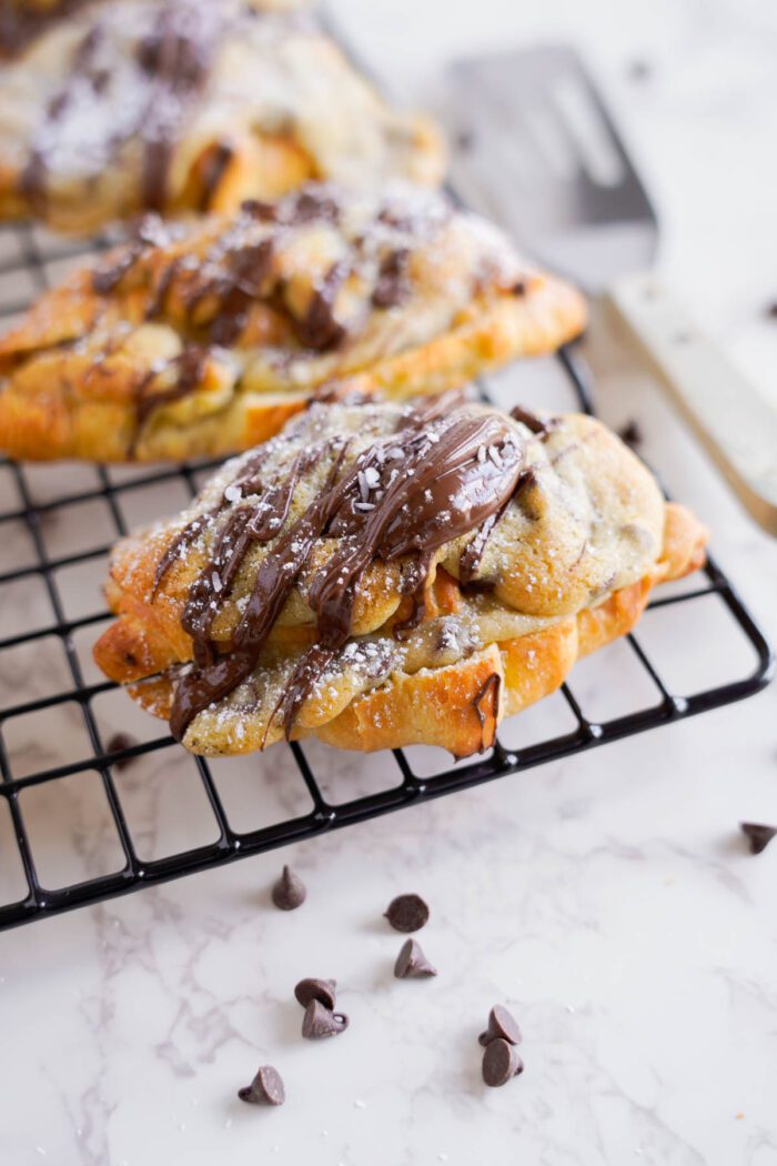 Freshly baked chocolate chip croissants on a cooling rack, sprinkled with powdered sugar and drizzled with chocolate, surrounded by chocolate chips.