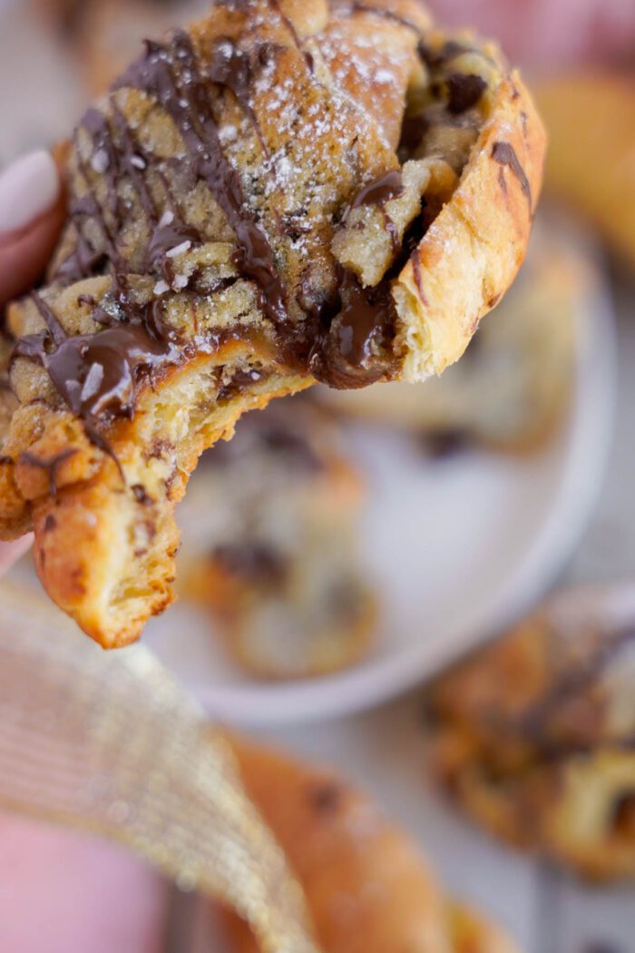 A close-up of a hand holding a chocolate chip cookie croissant with a bite taken out of it, over a plate of similar pastries.