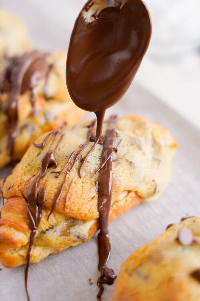 A spoon drizzling melted chocolate over freshly baked chocolate chip cookie croissants.