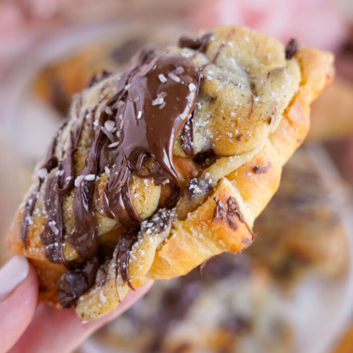 A close-up of a hand holding a chocolate-drizzled, freshly baked chocolate chip cookie with visible chunks of chocolate.