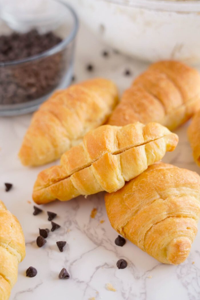 Freshly baked chocolate chip cookie croissants on a marble surface with chocolate chips scattered around and a bowl of chocolate chips in the background.