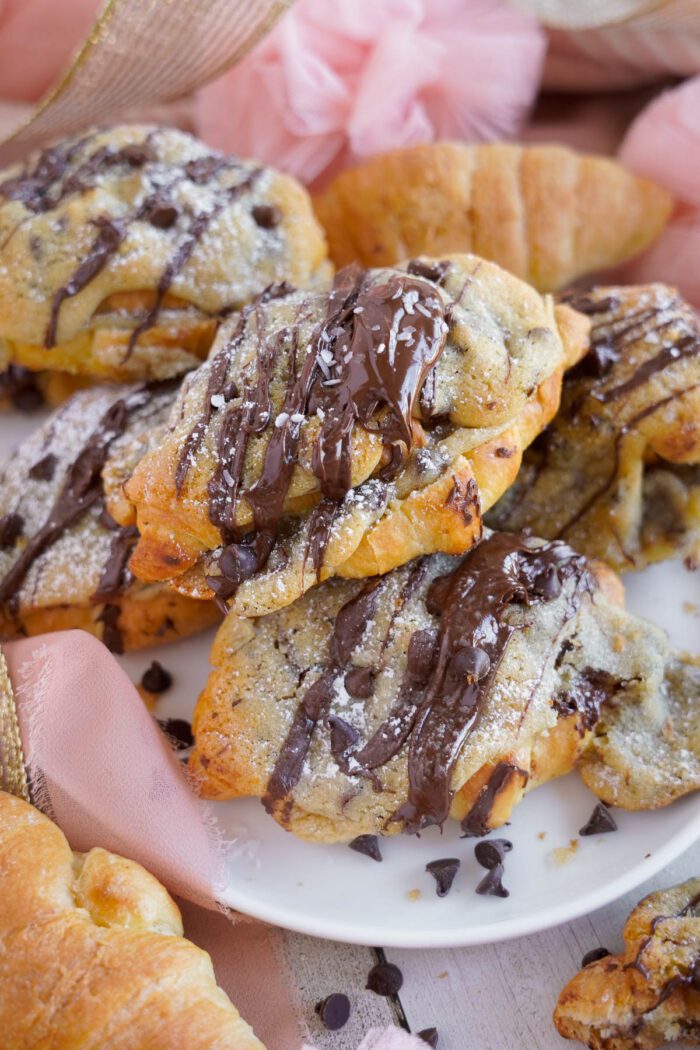 Plate of freshly baked chocolate chip cookie croissants topped with powdered sugar and drizzled with chocolate, surrounded by more pastries and pink fabric.