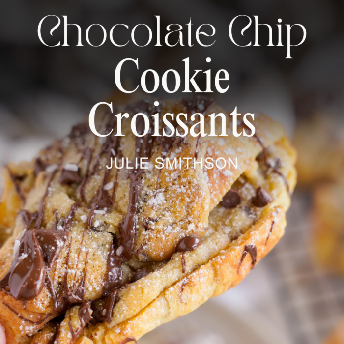 Close-up of a hand holding a chocolate chip cookie croissant, topped with chocolate drizzle and powdered sugar, with text "Chocolate Chip Cookie Croissant Recipe by Julie Smithson" above.