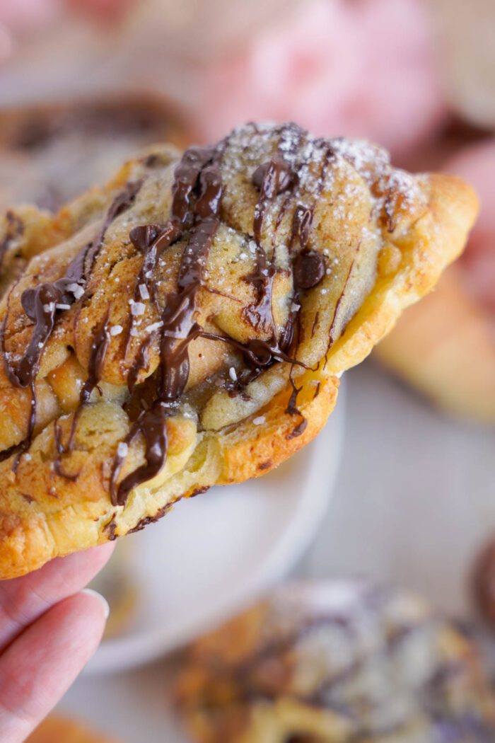 A hand holding a freshly baked Chocolate Chip Cookie Croissant drizzled with chocolate and sprinkled with powdered sugar, with more pastries blurred in the background.