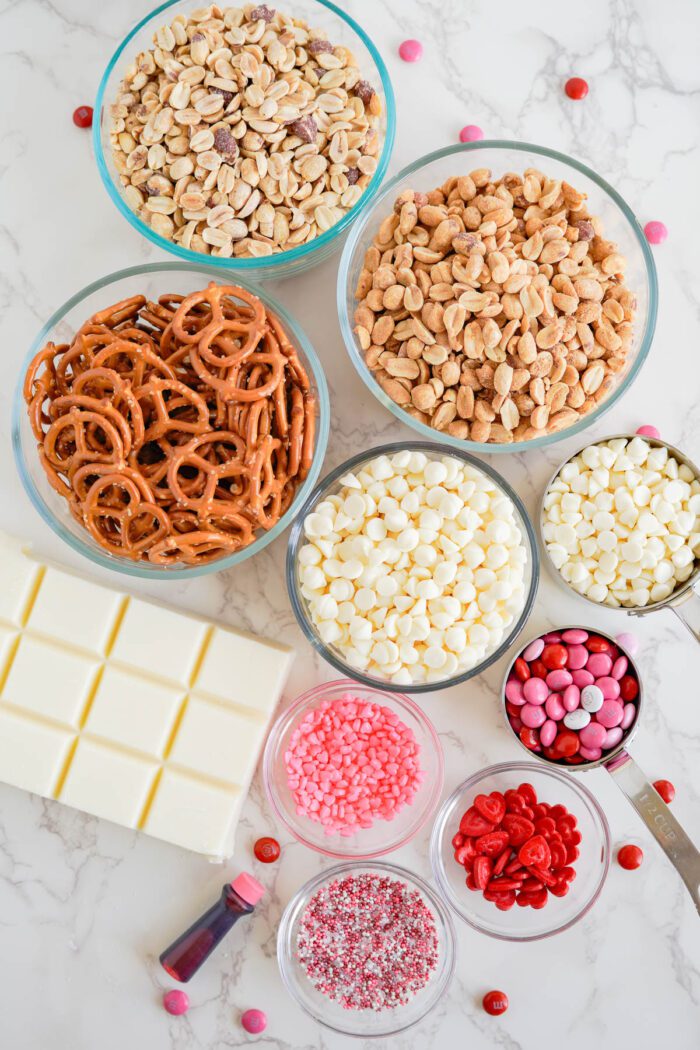 Various bowls of ingredients and sweets including pretzels, nuts, marshmallows, and candy, arranged with a chocolate bar and sprinkles on a marble surface.