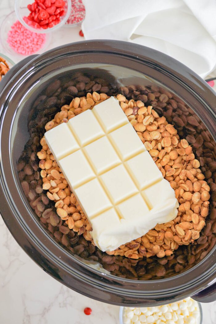 A slow cooker filled with peanuts, and a large block of white chocolate, with candy sprinkles visible in the background.