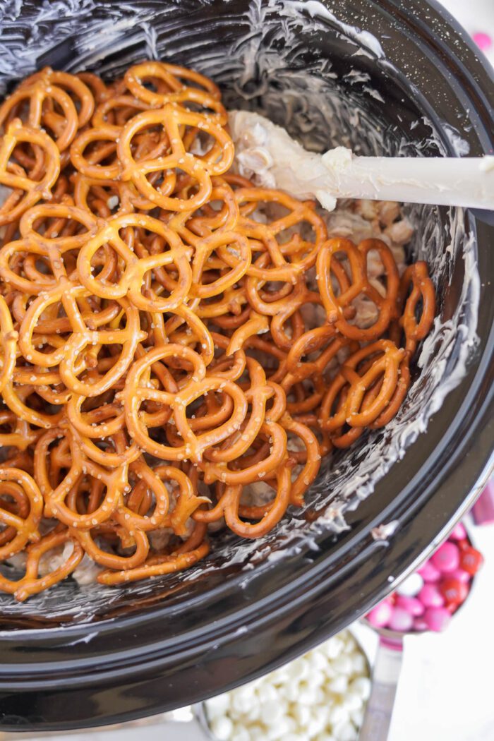 A crockpot containing pretzels mixed with white chocolate, with a spatula stirring the mixture.