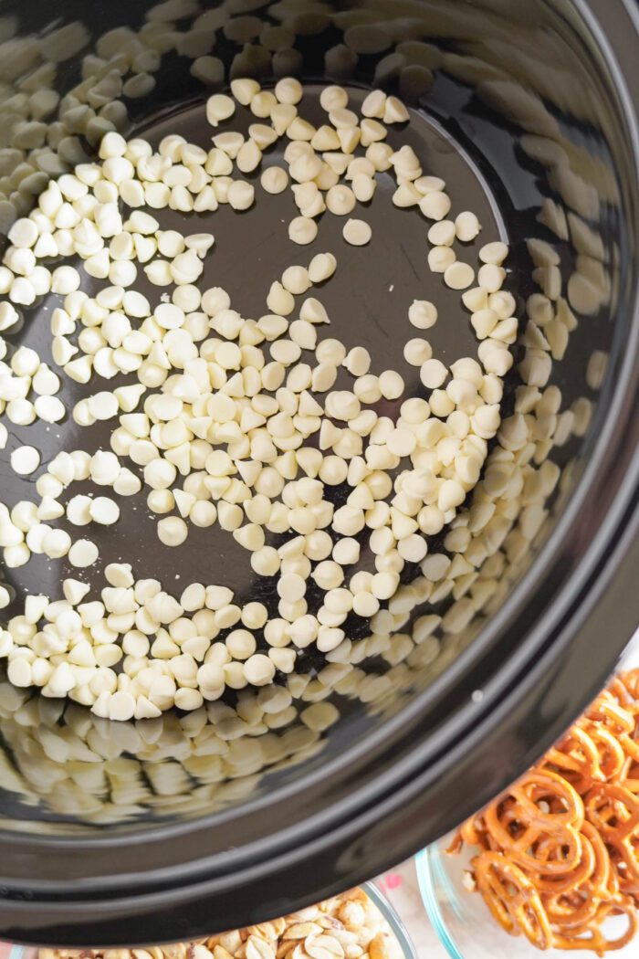 White chocolate chips in a crockpot, next to a bowl of pretzels and peanuts.