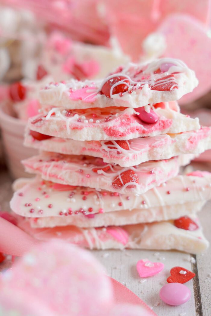 Stack of white chocolate bark with heart sprinkles and red candies on a wooden surface, with a pink bowl in the background.