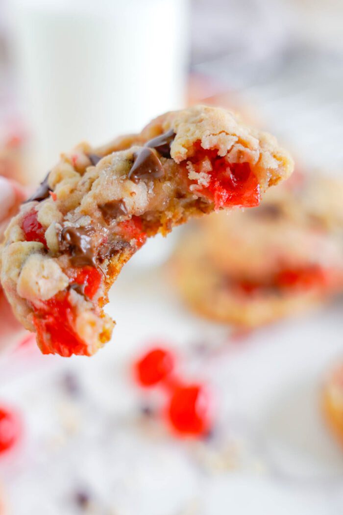 A close-up of a half-eaten cherry chocolate chip cookie with melting chocolate chips and bits of red fruit, alongside a glass of milk in the background.