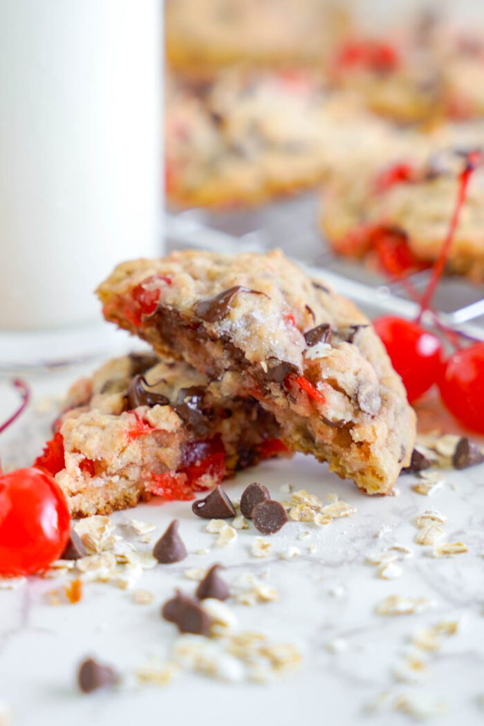 A close-up of a freshly baked Cherry Chocolate Chip Cookie with chocolate chips and cherries, broken in half to show the gooey interior, with a glass of milk and scattered ingredients in the background.
