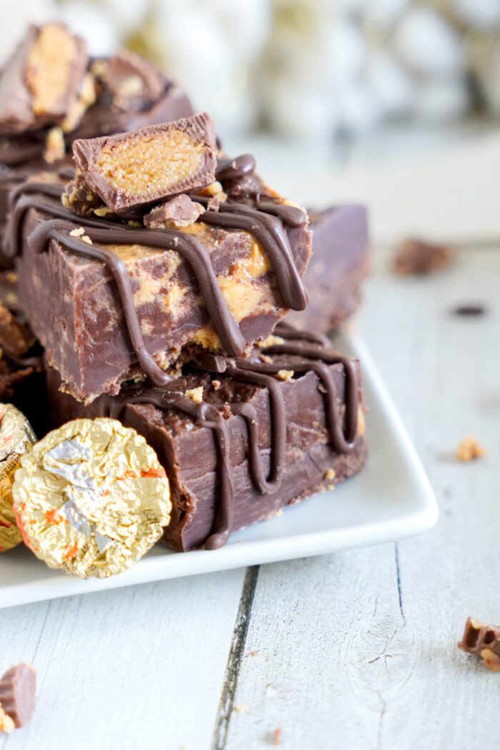 A stack of chocolate fudge with peanut butter cups and a drizzle of chocolate on a white plate, placed on a wooden surface.