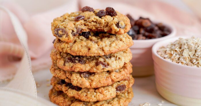 A stack of oatmeal raisin cookies on a light pink surface, with a bowl of oats and raisins in the background, showcasing the best oatmeal raisin cookies recipe.
