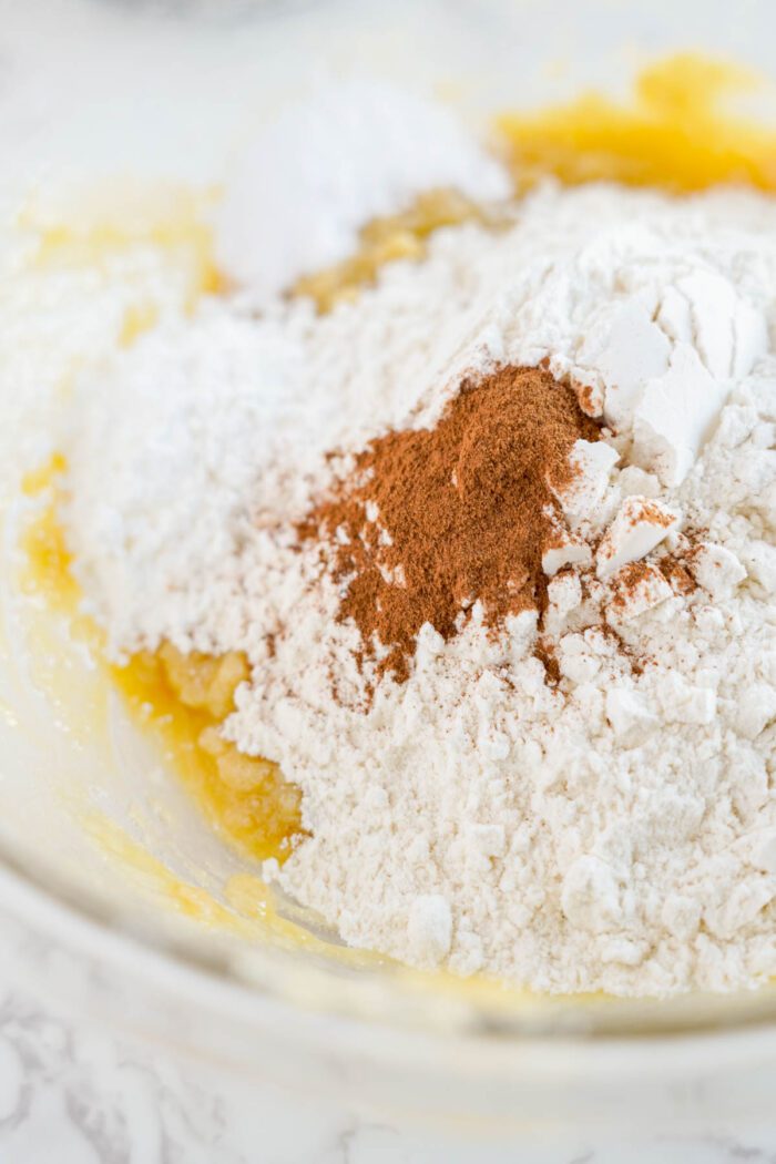 A glass bowl containing flour, a cracked egg, melted butter, oatmeal, raisins, and a sprinkle of cinnamon, ready for mixing.