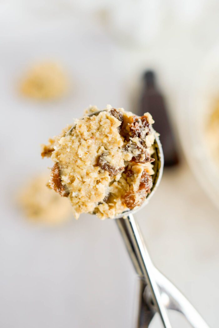 A close-up of a cookie dough scoop with raw oatmeal raisin dough, held above a bowl of the same mixture.