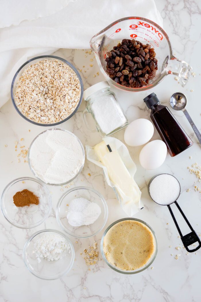 Ingredients for making the best oatmeal raisin cookies displayed on a marble countertop, including oats, flour, eggs, sugar, butter, and raisins.