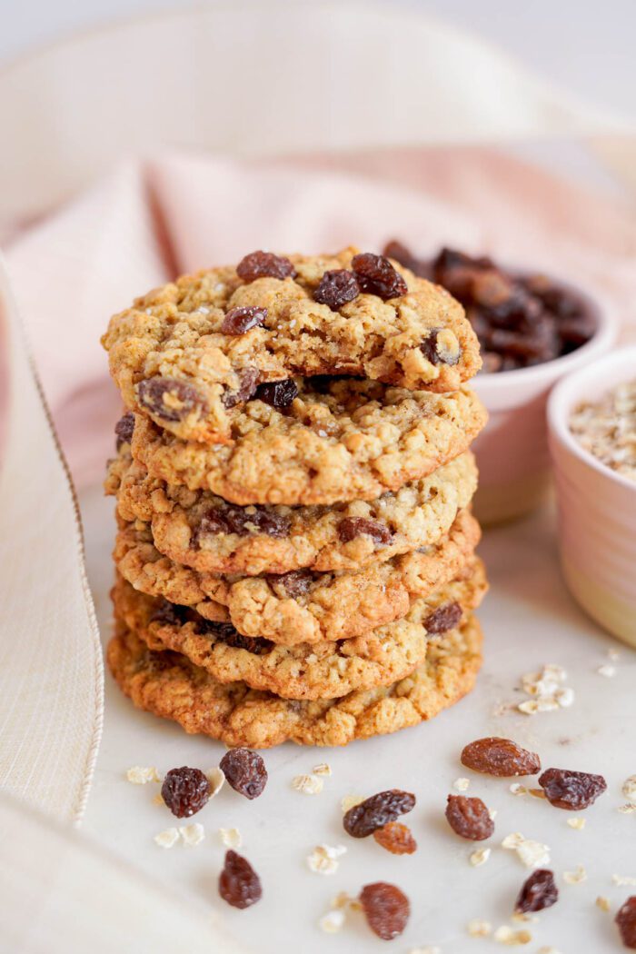 A stack of the best oatmeal raisin cookies on a white surface, with loose raisins and oats scattered around, and a cloth in the background.